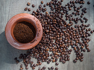 Naklejka premium Black coffee ground in a clay mortar and in grains on the surface of a wooden table in sunny rays. Top view.