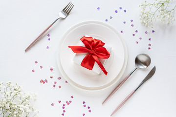 Flat lay overhead festive composition with gift box on the plate, cutlery set, beautiful flowers and decor on white background. Valentine’s day concept. table setting.