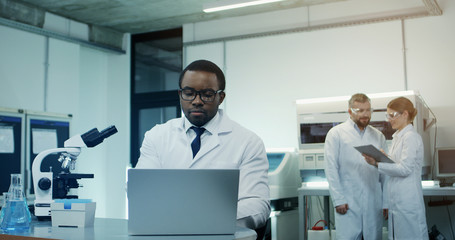 Naklejka na ściany i meble African American young man scientist in the white robe and glasses working at the laptop and microscope over research while his female caucasian co-worker coming with a tablet