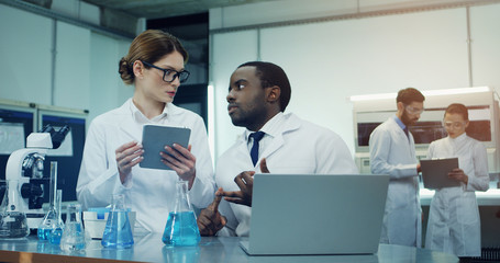 African American man and Caucasian woman working and talking in the laboratory while he sitting at...