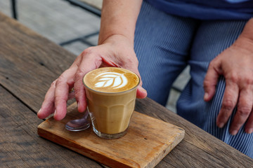 Selected focus view at Hhand of person pick and hold one cup of cappuccino or latte with flower pattern of latte art in transparent glass on wooden tray and table. Hipster vibes and vintage tone.