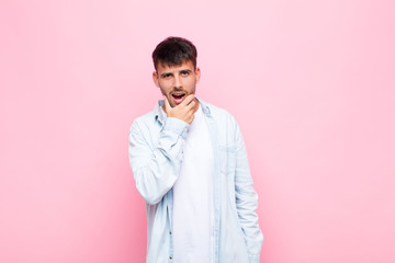 young handsome man with mouth and eyes wide open and hand on chin, feeling unpleasantly shocked,...