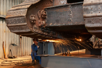 A worker performs grinding work on a old armored tank in a factory workshop.
