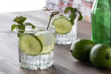 A close up of two glasses of sparkling water with slices of lime and mint on a wooden table against a bright window.