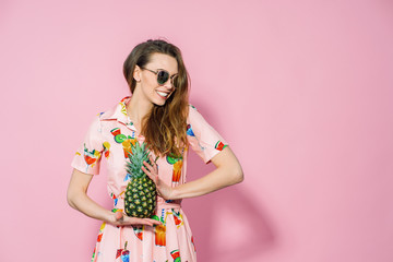 Beautiful woman in dress with printed friuts posing with pineapple on pink background. Pineapple in...