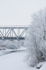 Winter landscape with railroad bridge over frozen river, snow-covered trees, hoarfrost.