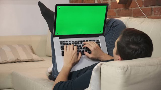 The person at the table is browsing the Internet on a green laptop screen. In a well-lit, cozy apartment. A man works in a loft office