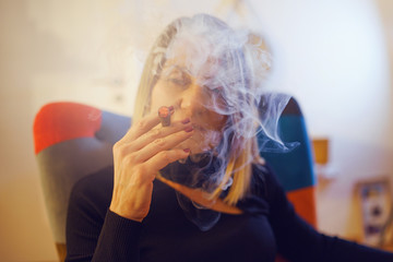 Caucasian mature woman in black lighting cigarillo at home while sitting in a colorful chair in the evening smoke smoking tobacco