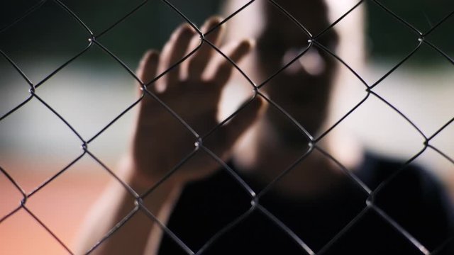 Scary shot of male zombie coming towards fence to attack. Horror scene. Caucasian imprizoned young man leaning on metal fence at basketball court at night.