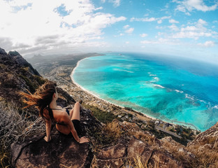Fototapeta na wymiar Woman sitting on top of rocky cliffs overlooking bright blue ocean, hair flowing in the wind