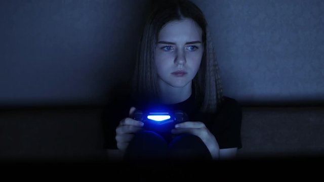 Frightened female teenager plays a scary game at night in the dark on a game console with a wireless joystick, at home