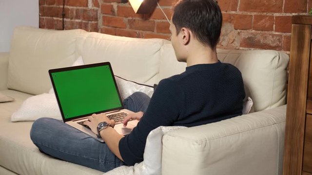 The person at the table is browsing the Internet on a green laptop screen. In a well-lit, cozy apartment. A man works in a loft office