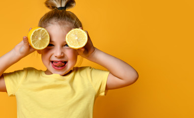 Little smiling cute blond girl in yellow t-shirt holding halves of fresh sour lemon fruit near eyes...