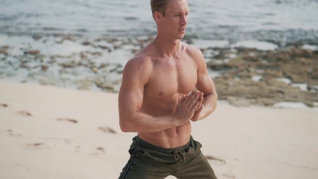 Man with muscular body doing yoga exercises on beach at sunset, slow motion.