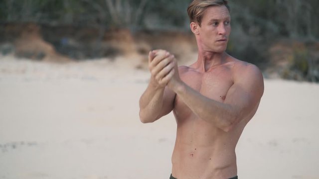 Man with muscular body doing streching exercises on beach at sunset, slow motion.