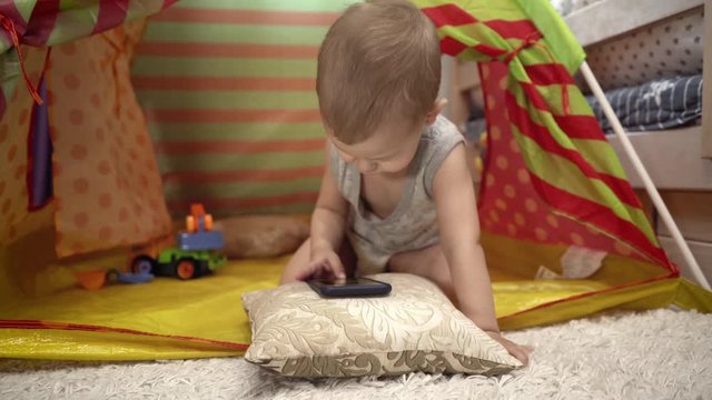 Charming baby plays with the phone in the children's tent. He watches videos and pictures.