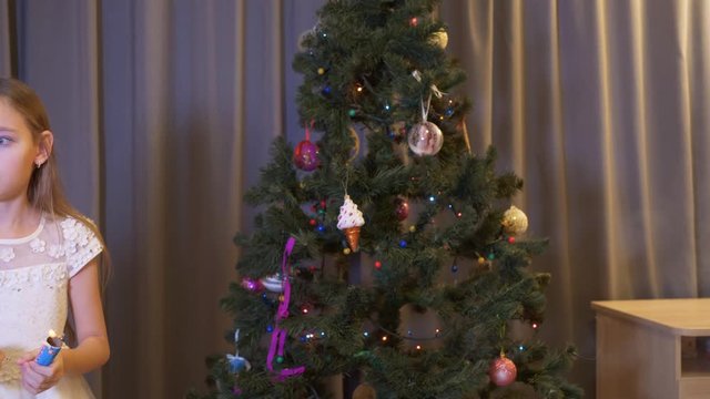 Adorable emotional little girl holding christmas cracker at home. Cute scared girl standing near christmas tree with shiny baubles and having fun with cracker at christmastime
