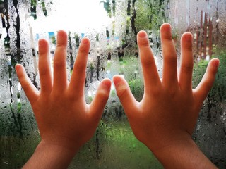 child's hand is placed on the window glass and there are water drops on the outside.