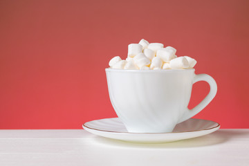 Small marshmallow slices in a white Cup on a white table on a pink background.