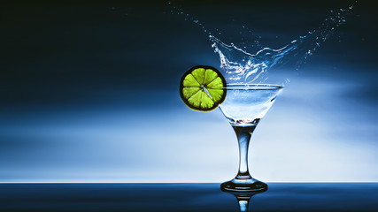 Alcohol cocktail with splash and green lime isolated on blue background. Vermouth cocktail inside martini glass over dark background. Copy space. Space for text.