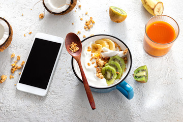 Breakfast table concept. Smartphone with сoconut yogurt with granola and fruits on light home table with with carrot apple juice