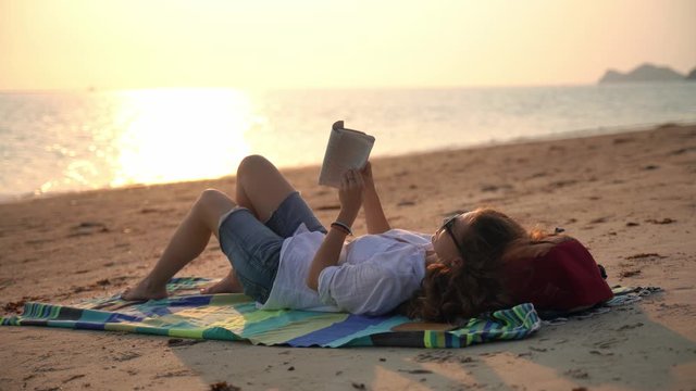 A young woman reading a book and relaxing on the beach at the sunset time.
