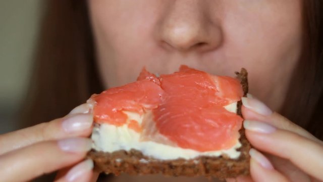 Closeup woman who eating a sandwich with red fish and butter. Girl mouth bites a salmon sandwich from whole wheat bread.