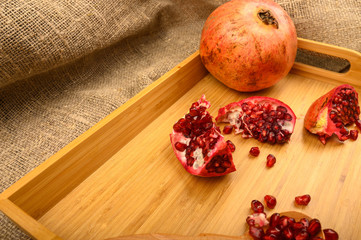 Ripe juicy pomegranate, pieces of pomegranate, pomegranate seeds on a wooden spoon on a wooden tray on a background of rough homespun fabric. Close up.