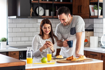 Couple in the kitchen on breakfast with orange juice and pastry on table looking on the mobile phone and smile