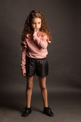 Studio portrait of a little girl, with curly curly hair, in pink blouse and black shorts, dancing on a dark red background