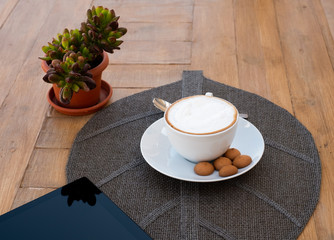 A wooden table prepared with a hot homemade cappuccino and something sweet.  One technological device as a  tablet to stay updated