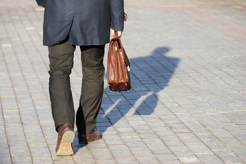 Man in a business suit carrying leather briefcase walking on the street, shadow on a pavement....