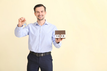 Male real estate agent with key and model of house on light background