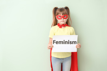 Little girl in superhero costume holding paper with word FEMINISM on color background