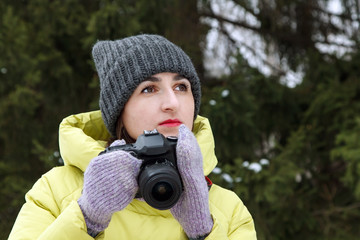 Young woman with a camera in a winter park on a background of fir trees. A girl photographer holds a camera in hand. Copy space