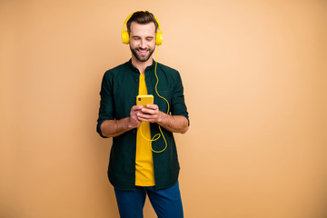 Photo of attractive funny guy holding telephone hands listening youth songs cool modern yellow earphones wear casual clothes isolated beige color background