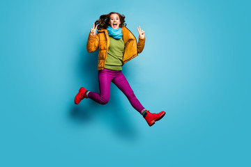 Fototapeta na wymiar Full length photo of pretty lady jump high up showing v-sign symbols wear casual yellow overcoat scarf violet pants red boots green pullover isolated blue color background