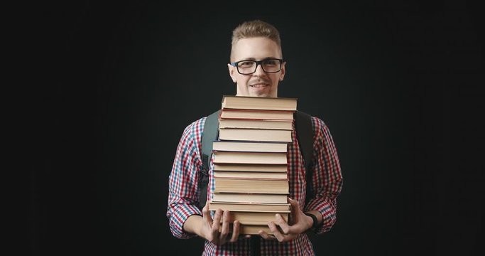 Young male student in eyeglasses holding heavy stack of books and looking to camera, nerd