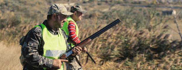 A mans with a gun in his hands and an camouflage vest on a pheasant hunt in a wooded area in cloudy...