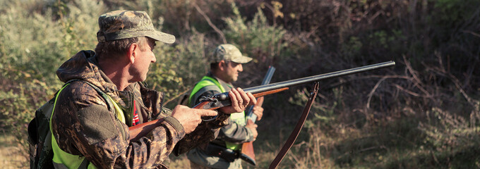 A mans with a gun in his hands and an camouflage vest on a pheasant hunt in a wooded area in cloudy...