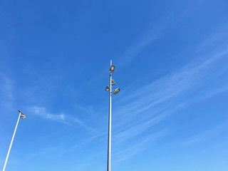 Tall metal poles with street lights are seen from below. One pole has four lights, the other one. The sky is blue with trails of faint white clouds.