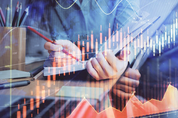 A woman hands writing information about stock market in notepad. Forex chart holograms in front. Concept of research. Multi exposure