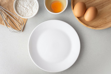 Top view above getting ready to bake, whisk, bowl of flour, small bowl of cracked egg white egg yolk, and raw eggs with empty white plate on grey background.
