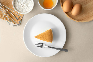 Top view above piece egg cake with fork in a white plate with ingredients such as whisk, bowl of flour, small bowl of cracked egg white egg yolk, and raw eggs on grey background.