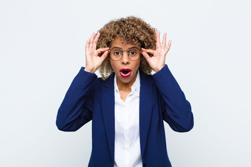 young woman african american feeling shocked, amazed and surprised, holding glasses with astonished, disbelieving look against flat wall - obrazy, fototapety, plakaty