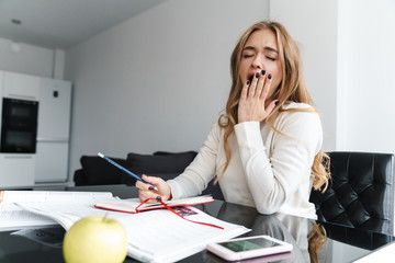 Photo of young sleepy woman yawning and making notes in exercise book - obrazy, fototapety, plakaty