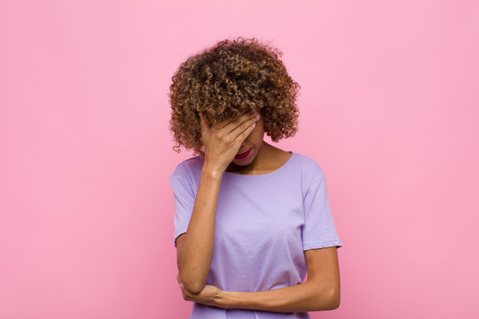 young african american woman looking stressed, ashamed or upset, with a headache, covering face with hand against pink wall