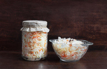 Homemade sauerkraut with carrots in a glass jar and bowl on a dark wooden background. Fermented food. Marinated vegetables