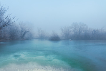 Frozen turquoise lake in misty winter morning, with trees without leaves on a horizon 