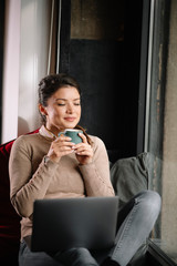 Young businesswoman working from home. Beautiful woman sitting on window and using laptop. 